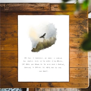 May include: A framed art print featuring a watercolor-style image of a bird in flight above a landscape. The print includes a quote by Anne Brontë in a handwritten font. The artwork is displayed on a wooden table.
