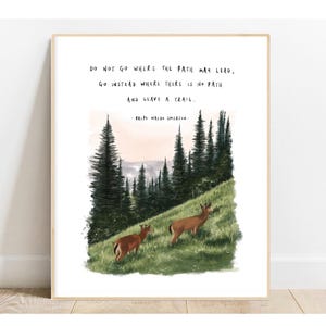 May include: A framed art print featuring a watercolor illustration of two deer on a grassy hillside with evergreen trees. The print includes the quote "DO NOT GO WHERE THE PATH MAY LEAD, GO INSTEAD WHERE THERE IS NO PATH AND LEAVE A TRAIL." by Ralph Waldo Emerson.