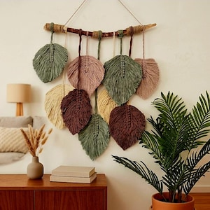 May include: A macrame wall hanging featuring multiple leaf-shaped fiber elements in shades of sage green, dusty pink, cream, and dark brown. The leaves are suspended from a horizontal wooden branch wrapped with twine and hung by a white cord.