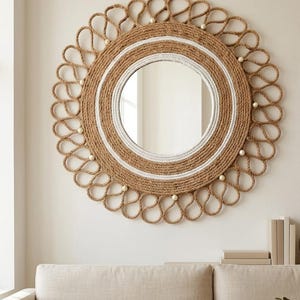 Könnte beinhalten: Runder Wandspiegel mit einem Design aus Naturseil. Der Spiegel hat einen gewebten Seilrahmen mit einem weißen Streifen und dekorativen Perlen. Er hängt an einer neutralen Wand über einem beigen Sofa.