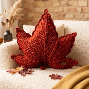 May include: A rust-colored, leaf-shaped crochet pillow with a textured, ribbed surface and a small brown oval tag attached to the lower right side. The pillow features a central vein detail and pointed lobes, mimicking the appearance of a maple leaf.