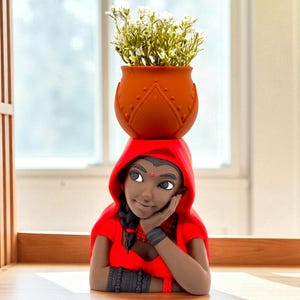 May include: A terracotta-colored flower pot with a lotus design, balanced on the head of a figurine wearing a red hooded cloak. The figurine has dark skin, a bindi, and is resting her chin on her hand. The pot contains small white flowers.