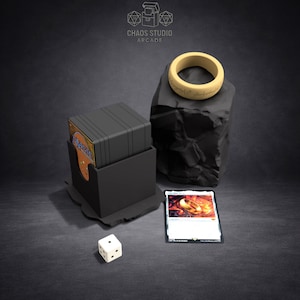 May include: A product shot featuring a black card holder with a deck of cards, a white die, and a Magic: The Gathering card. A gold ring with engraved text sits atop a black rock. The Chaos Studio Arcade logo is in the top left.