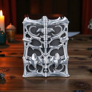 May include: A silver-colored, rectangular box with an ornate, gothic design. The box features a central spine-like detail and decorative, pointed edges. The box is made of metal and sits on a wooden surface.
