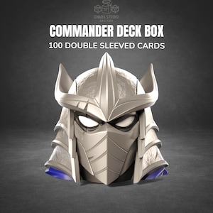 May include: A Commander Deck Box with a silver Shredder helmet design, holding 100 double-sleeved cards. The box has a blue base and the text "COMMANDER DECK BOX" and "100 DOUBLE SLEEVED CARDS" in white.