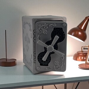 May include: A gray, decorative safe box with ornate floral and scrollwork details. The safe features a combination dial and a hinged door. The box is sitting on a white surface.
