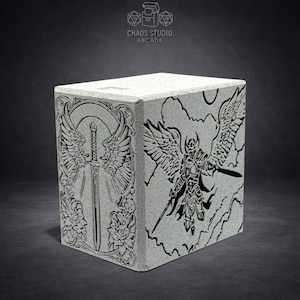 May include: A gray, textured box with engraved artwork. One side features a sword with wings and floral accents. The other side depicts an armored figure with wings and a sword. The top of the box is plain. The text "CHAOS STUDIO ARCADE" is visible.
