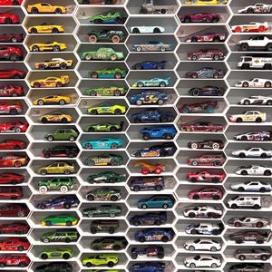 May include: A collection of colorful toy cars displayed in a honeycomb-shaped shelf. The cars are various colors, including red, yellow, green, blue, and white. Some cars have racing stripes and other decorative details. The shelf is white.