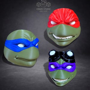 May include: Three Teenage Mutant Ninja Turtle masks in green, red, blue, and purple. The masks have white eye coverings and are smiling. The Chaos Studio Arcade logo is in the top left corner.