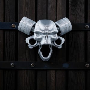 May include: A silver-colored skull-shaped door knocker with an open mouth and two barbell-shaped handles. The knocker is mounted on a dark wooden door with black metal accents.