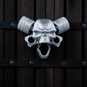May include: A silver-colored skull-shaped door knocker with a wide open mouth and two barbell-shaped handles. The knocker is mounted on a dark wooden door with black metal accents.
