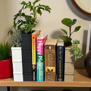 May include: A shelf displays a collection of books with plants in decorative pots. The books include titles like "Happy Place" and "Verity." The pots are white, black, and beige, holding various succulents and leafy plants. A small bunny book mark is in one of the books.