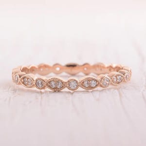 May include: Rose gold ring with a repeating pattern of marquise and round-cut clear gemstones. The ring has a delicate, ornate design, perfect for everyday wear or special occasions. The ring is set on a white surface.