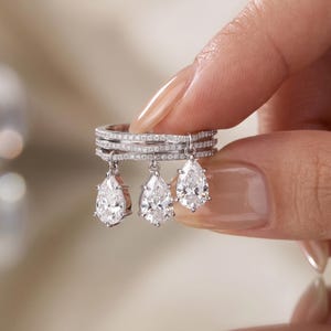 Op de afbeelding: Een zilverkleurige ring met drie gestapelde banden bezet met kleine heldere steentjes. Drie peervormige heldere stenen hangen aan de banden, elk bevestigd door een klein metalen lusje, wat een bungelend effect creëert aan de voorzijde.