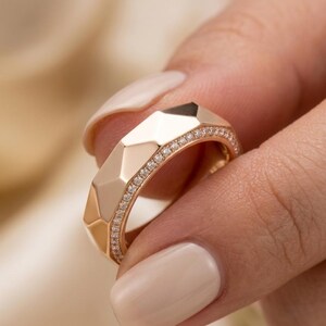 May include: A gold ring with a geometric design. The ring features a series of angled facets and is lined with small, sparkling stones. The ring is held up by a person's fingers.