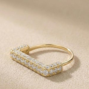 May include: A gold-colored ring with a rectangular design, encrusted with small, clear gemstones. The ring has a smooth, polished band and a geometric shape, showcasing a modern jewelry style.
