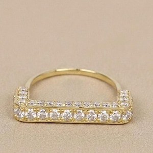 May include: A gold-colored ring with a rectangular face, encrusted with multiple small, clear gemstones. The band is a simple, rounded design. The ring is set against a neutral background, highlighting its sparkle and design.