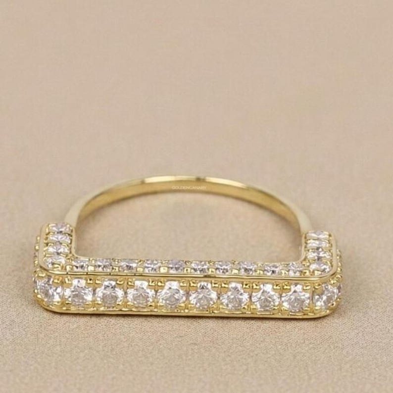 May include: A gold-colored ring with a rectangular face, encrusted with multiple small, clear gemstones. The band is a simple, rounded design. The ring is set against a neutral background, highlighting its sparkle and design.