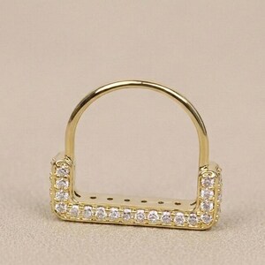 May include: A gold-colored ring with a rectangular base and a curved top. The rectangular portion is encrusted with small, clear gemstones. The ring is set against a neutral background, highlighting its design and sparkle.
