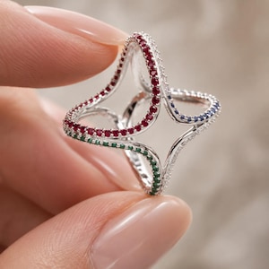 May include: A silver-toned ring with a unique, openwork design. The ring is adorned with small, colorful gemstones in red, green, blue, and clear. The gemstones are set along the edges of the ring, creating a sparkling effect.