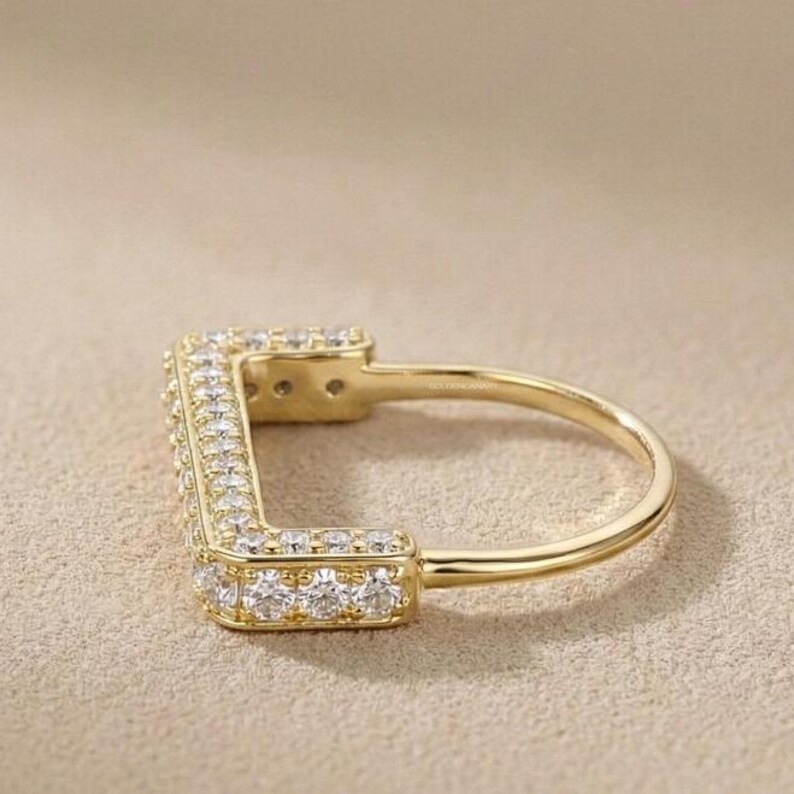 May include: A gold-colored ring with a square design, encrusted with small, clear gemstones. The ring has a smooth, polished band and a rectangular top lined with the gemstones. The ring is set against a neutral background.