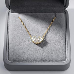 May include: A gold necklace with a pendant of two heart-shaped diamonds. The necklace is presented in a grey jewellery box with a velvet lining. The diamonds sparkle, reflecting light.