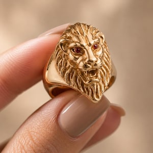 Könnte beinhalten: Ein goldfarbener Ring mit einem detaillierten Löwenkopf und roten Edelsteinaugen. Der Ring hat eine strukturierte Mähne. Nahaufnahme, die das Design und die Handwerkskunst des Schmucks zeigt.