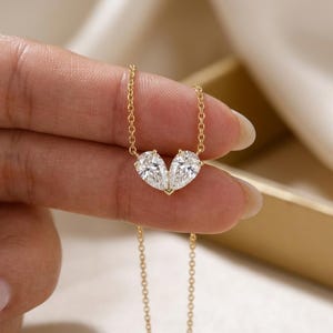 May include: A gold necklace with two pear-cut, clear gemstones arranged to form a heart. The gemstones are linked by a fine gold chain. The necklace is shown against a soft, neutral backdrop. The gemstones are likely to be measured in carats.