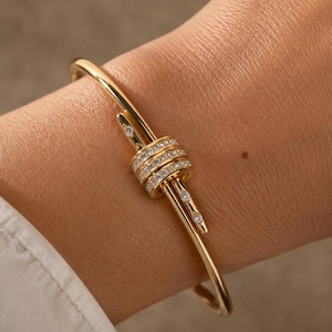May include: A gold bangle bracelet featuring a central knot design composed of three stacked rings encrusted with small clear stones. The two ends of the bangle extend from the knot, each finished with a single small clear stone at the tip.