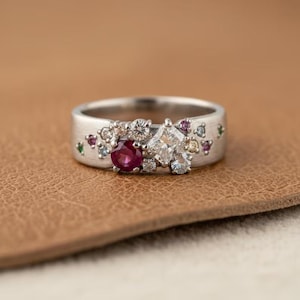 May include: A silver ring with a cluster of gemstones. The ring features a large, square-cut diamond, a round ruby, and several smaller diamonds and colored gemstones. The ring is set on a textured band.
