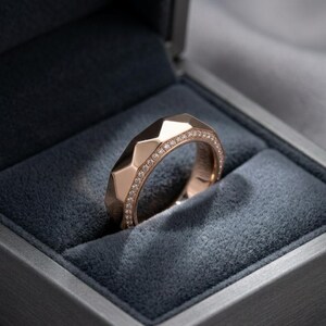 May include: A rose gold ring with a geometric design, set with small diamonds along the top edge. The ring is displayed inside a gray velvet-lined box, creating a luxurious presentation. The ring's faceted surface catches the light.