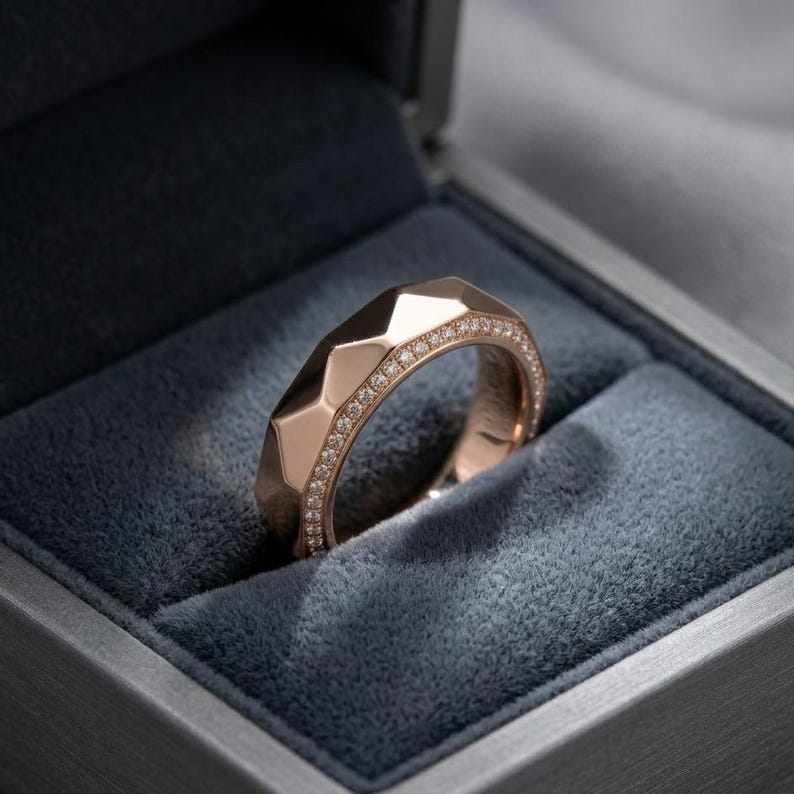 May include: A rose gold ring with a geometric design, set with small diamonds along the top edge. The ring is displayed inside a gray velvet-lined box, creating a luxurious presentation. The ring's faceted surface catches the light.