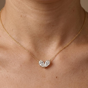 May include: A gold necklace with a pendant of two pear-cut, clear gemstones arranged to form a heart. The chain is visible around the neck. The gemstones are set close together.