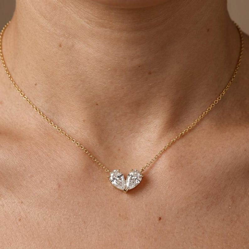 May include: A gold necklace with a pendant of two pear-cut, clear gemstones arranged to form a heart. The chain is visible around the neck. The gemstones are set close together.