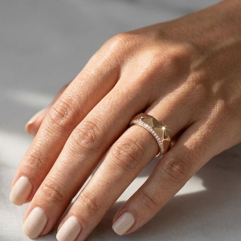 May include: A rose gold ring with a geometric design. The ring features a series of triangular facets and a band lined with small, sparkling stones. The ring is shown on a hand with polished nails.