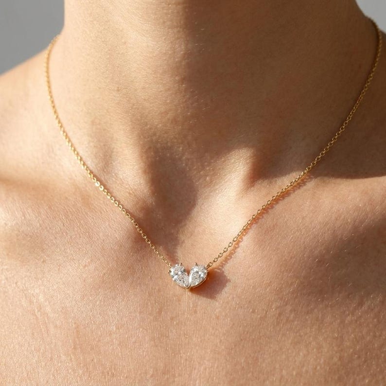 May include: A gold necklace with a pendant of two clear gemstones. The pendant is in the shape of a heart. The necklace is worn around the neck, highlighting the jewellery's design. The chain is thin and appears to be gold.