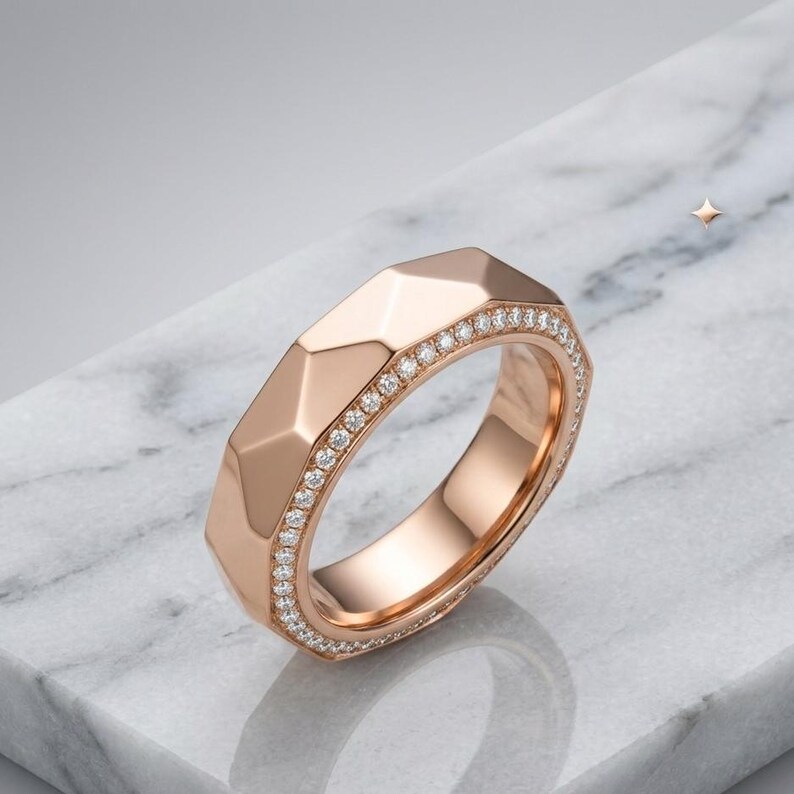 May include: Rose gold ring with a geometric design and a band of small, sparkling diamonds. The ring has a faceted surface, catching the light and creating a brilliant display. The ring is set against a marble surface.