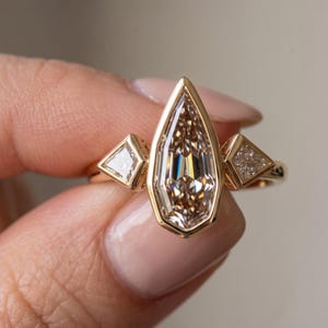 May include: A gold ring featuring a large, central, shield-shaped faceted stone in a bezel setting, flanked by two smaller, triangular-cut stones on either side. The band is crafted from polished yellow gold, creating a cohesive and elegant design.