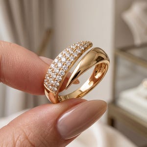 May include: A gold bypass ring featuring a polished, smooth band on one side and a tapered section encrusted with multiple small, clear, round-cut stones on the other. The ring has an open design where the two ends overlap slightly.