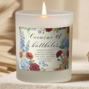 May include: A lit, frosted glass candle with a floral label that reads "Cocaine & Buttholes." The candle sits on a stone block, with dried plants in the background. The candle's flame is lit.