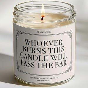 May include: A clear glass jar candle with a lit wick and a white label featuring the text "WHOEVER BURNS THIS CANDLE WILL PASS THE BAR" in black serif font. The label also includes "MUGNIFIQ USA" and "ECO FRIENDLY | VEGAN | MADE IN USA | NATURAL SOY WAX".