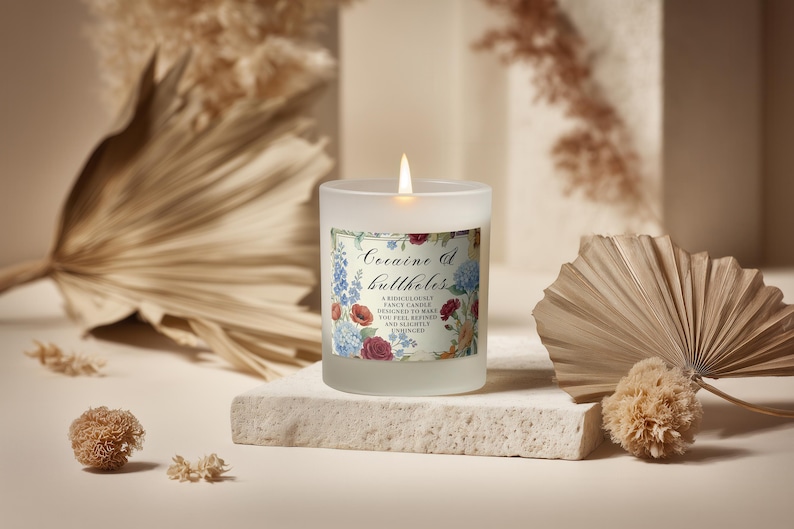 May include: A lit, frosted glass candle with a floral label that reads "Cocaine & Buttholes." The candle sits on a stone block, with dried plants in the background. The candle's flame is lit.