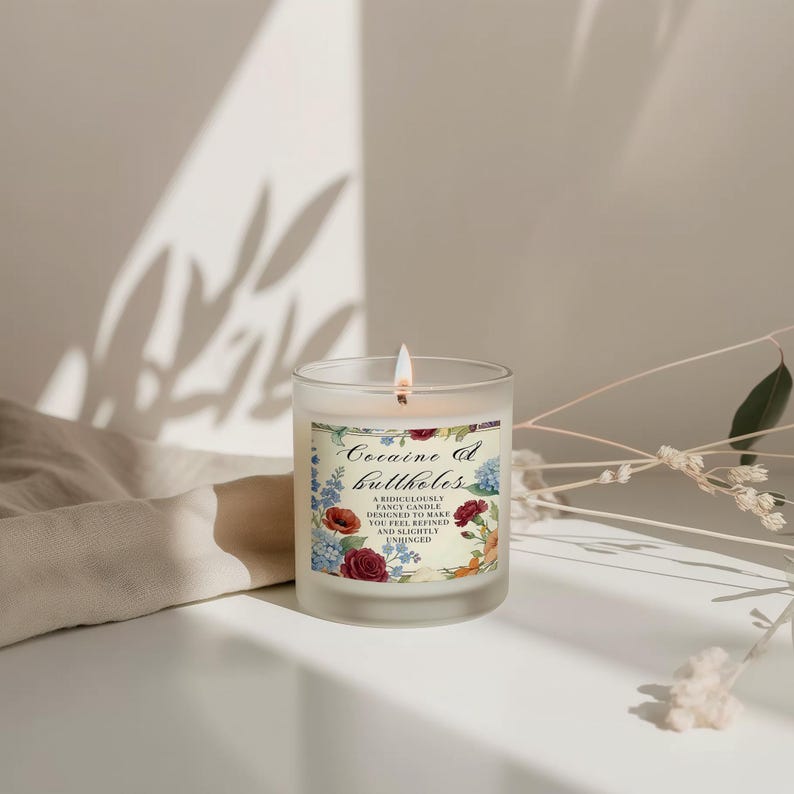 May include: A lit, frosted glass candle with a floral label that reads "Cocaine & Buttholes". The label features red poppies, blue hydrangeas, and other flowers. The candle is on a white surface.