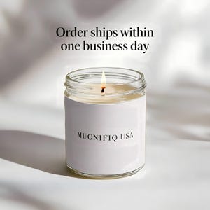 May include: A burning candle in a clear glass jar with a white label displaying "MUGNIFIQ USA". The text "Order ships within one business day" is above the jar. The candle sits on a white surface.