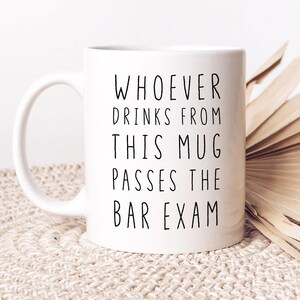 May include: White ceramic mug with a curved handle. The mug features black text that reads: "WHOEVER DRINKS FROM THIS MUG PASSES THE BAR EXAM."