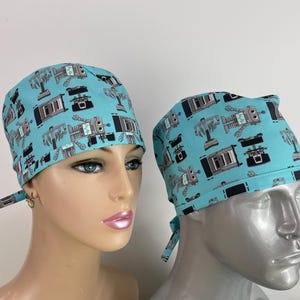 May include: Two turquoise blue surgical scrub hats with a pattern of robots. The hats have black ties.