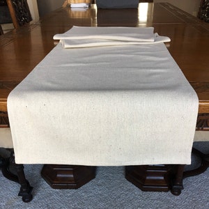 May include: A natural linen table runner in a light beige color. The runner is folded in half and placed on a wooden table.