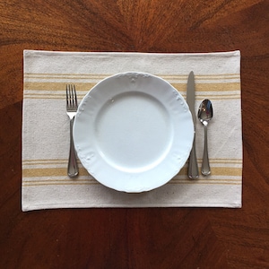 May include: A white dinner plate with a simple design sits on a beige placemat with yellow stripes. A fork, knife, and spoon are placed to the side of the plate.