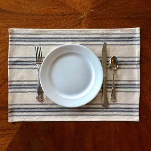 May include: A place setting on a wooden table. The setting includes a white plate, a fork, a knife, and a spoon. The plate rests on a rectangular placemat with blue and white stripes.