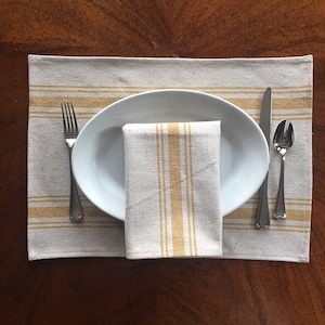 May include: A white oval plate with a folded linen napkin with yellow stripes sits on a beige placemat with yellow stripes. A fork, knife, and spoon are placed to the right of the plate.